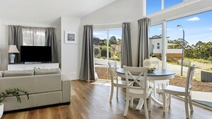 A modern living room with a flat-screen TV, a sofa, a dining table with chairs, and large windows offering a view of the outdoors.