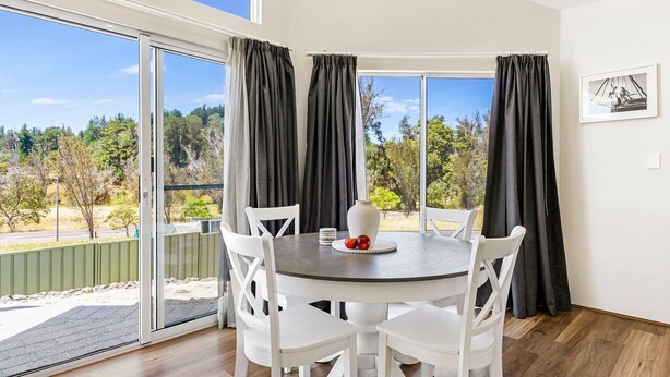 A dining area with a round table and white chairs, large windows with dark curtains, and a view of trees and a road.