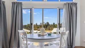 A round table with white chairs, a vase, and a bowl, set against a window with grey curtains.