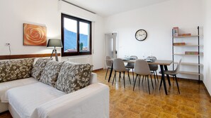 Una sala de estar con un sofá, una mesa de comedor con sillas, una estantería, un reloj y un ventanal con vistas.