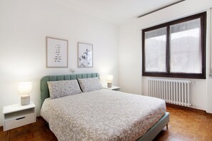 A bedroom with a bed, two bedside tables, a radiator, and a window with curtains.