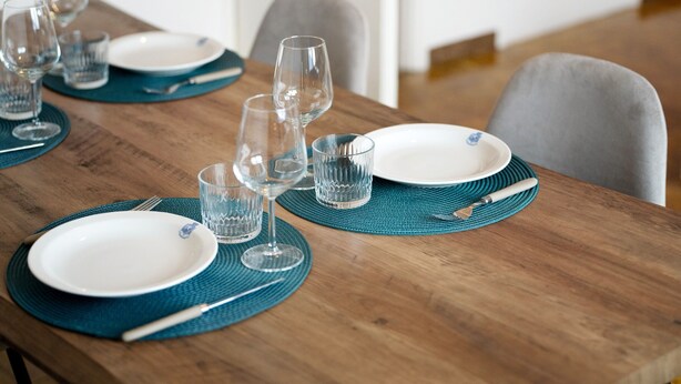 Una mesa de comedor dispuesta con platos, vasos y cubiertos, con superficie de madera y posavasos color turquesa.
