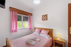 Ein Schlafzimmer mit einem Bett, einem Nachttisch mit Lampe, einem Fenster mit Vorhängen und einem an der Wand befestigten Kunstwerk.