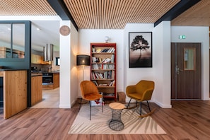 Un intérieur moderne avec un plafond en bois, une bibliothèque, deux chaises, une table ronde et un tableau encadré au mur.