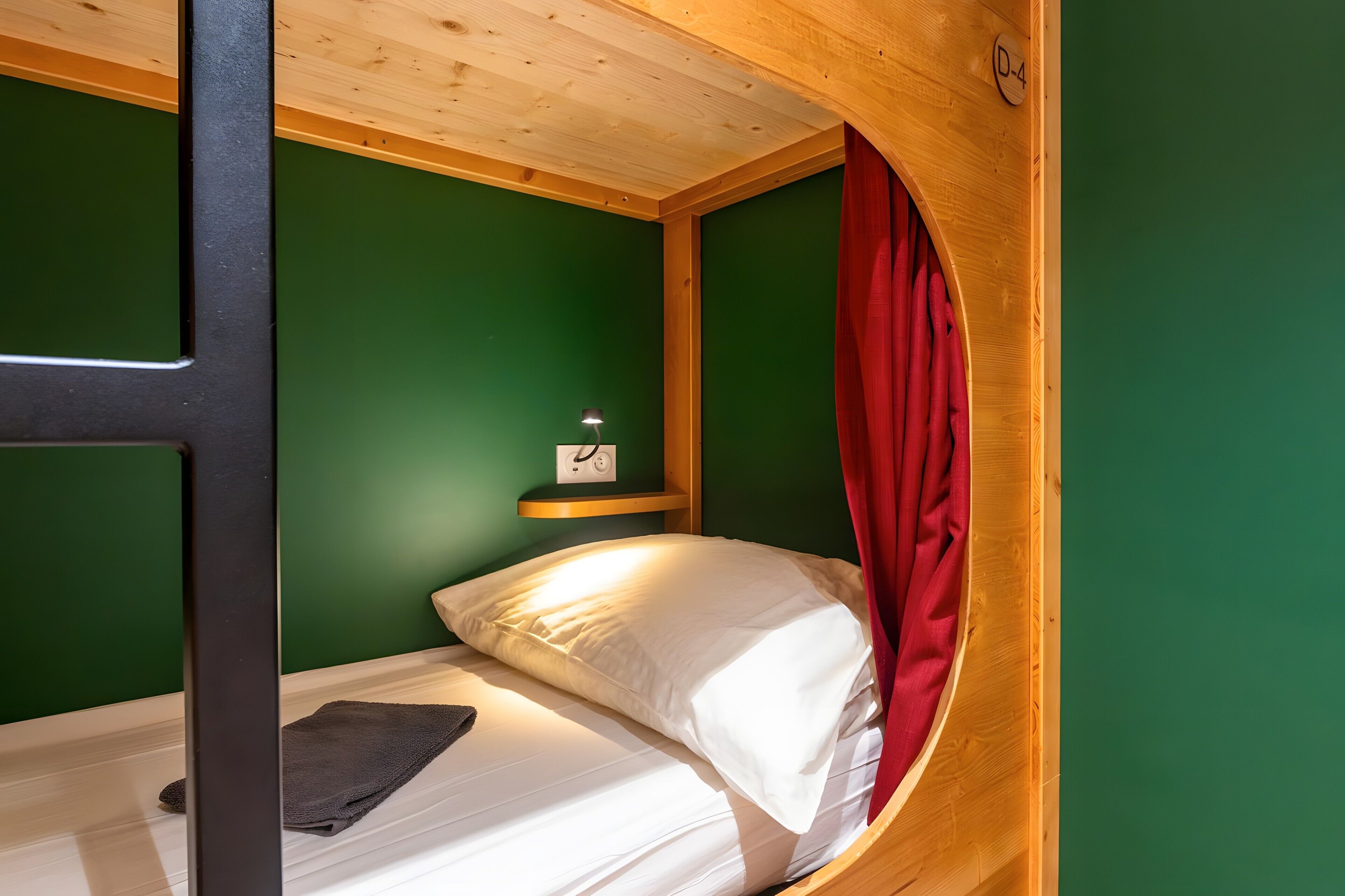 Un lit superposé avec une tête de lit en bois, un rideau rouge, une table de chevet avec une lampe et une horloge au mur.