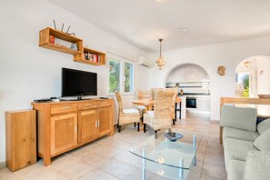 A modern living room with a TV, wooden cabinets, wicker chairs, a glass coffee table, and a kitchen in the background.