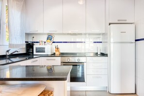 A modern kitchen with white cabinets, a black countertop, a refrigerator, a microwave, a stove, and a window with a striped curtain.