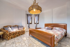 Ein Schlafzimmer mit einem hölzernen Bett, einem blumigen Sofa, einer gewebten Leuchte und einem Boden mit Steinplatten.
