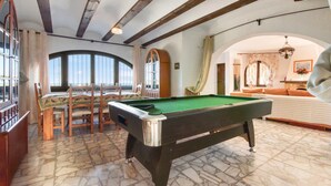 Una mesa de billar en una sala con techo alto y vigas a la vista, junto a un espacio de descanso con sofá y sillones.
