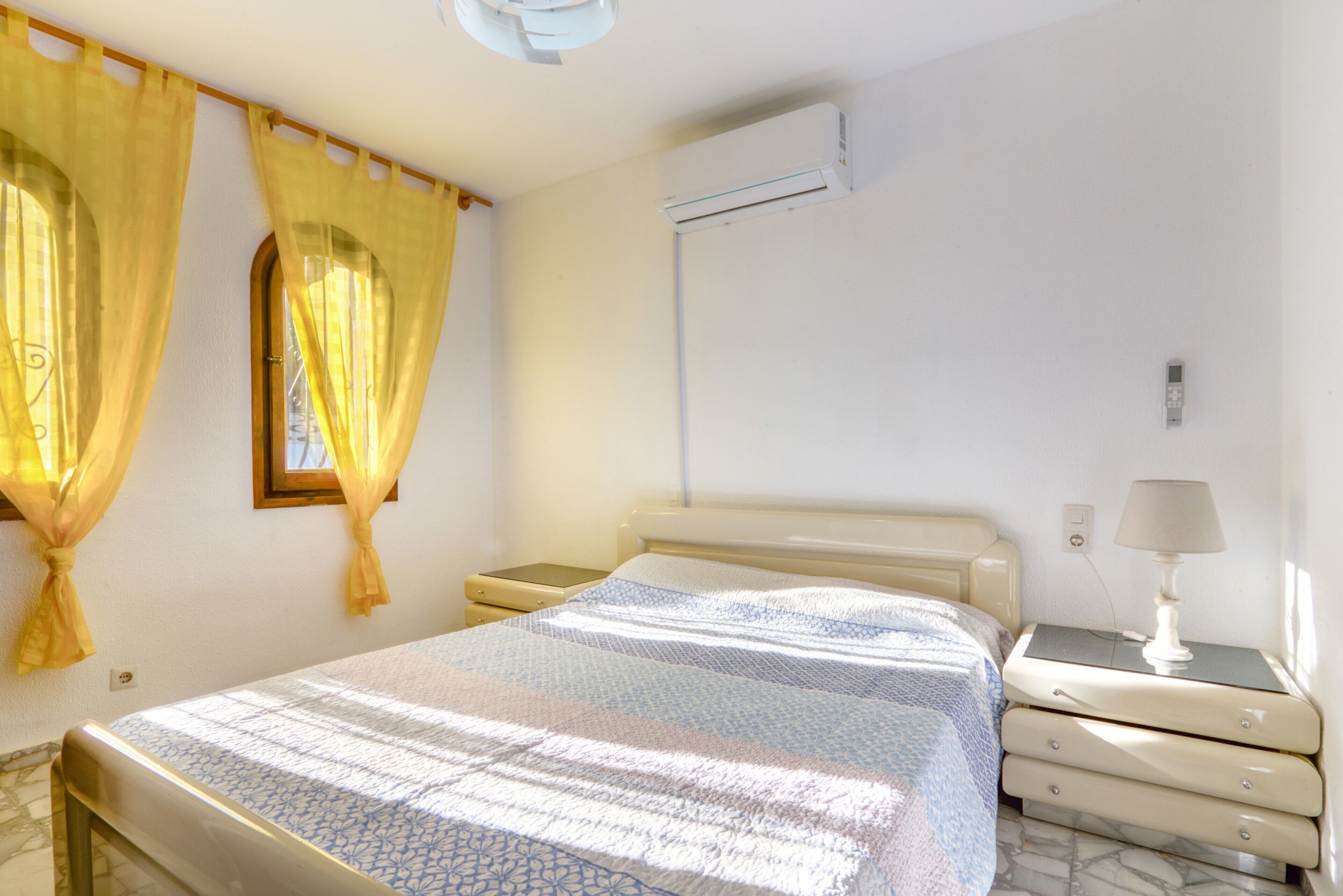 A bedroom with a bed, bedside tables, an air conditioning unit, curtains, and a lamp.