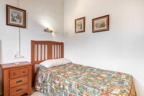 Ein Schlafzimmer mit einem hölzernen Kopfteil, einem Bett mit farbenfroher Steudecke, einem Nachttisch mit Schubladen und zwei gerahmten Bildern an der Wand.