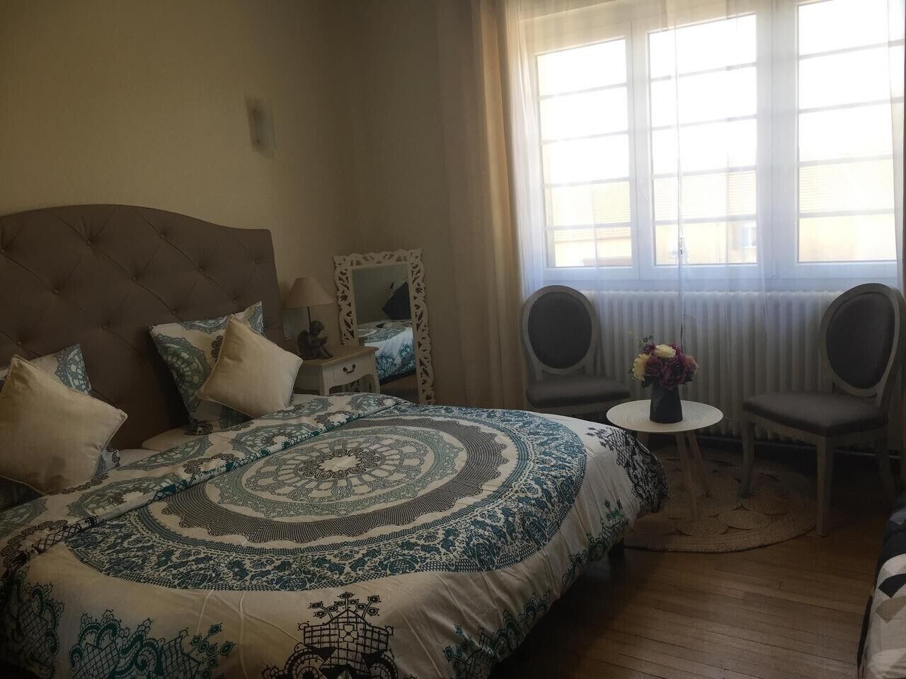 Een slaapkamer met een groot bed, twee stoelen, een tafeltje met bloemen en een raam met jaloezieën.