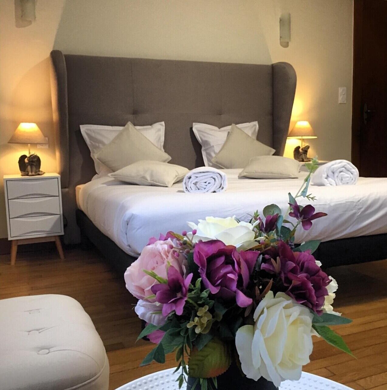 A hotel room with a bed, pillows, a nightstand, a lamp, a cat figurine, and a flower arrangement.