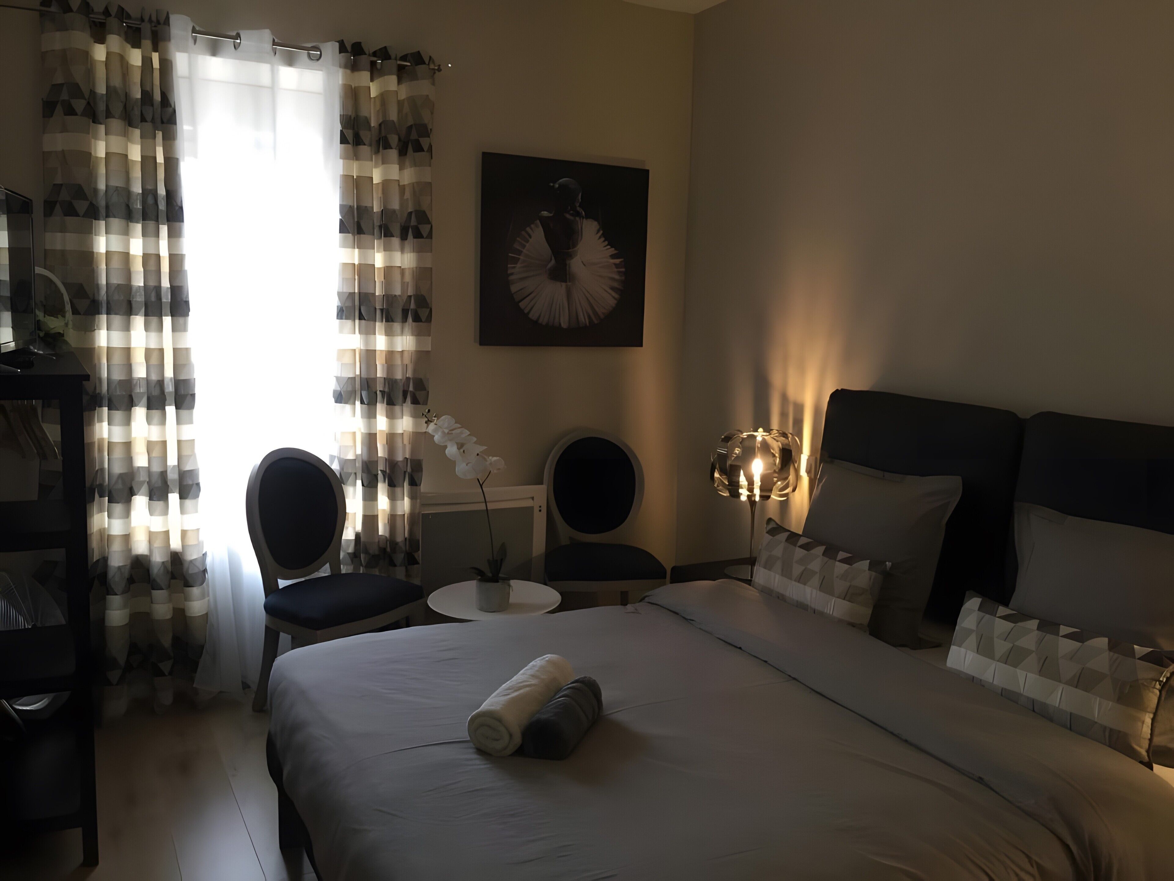 Une chambre d’hôtel avec un lit, deux chaises, une petite table, une lampe et un tableau au mur.