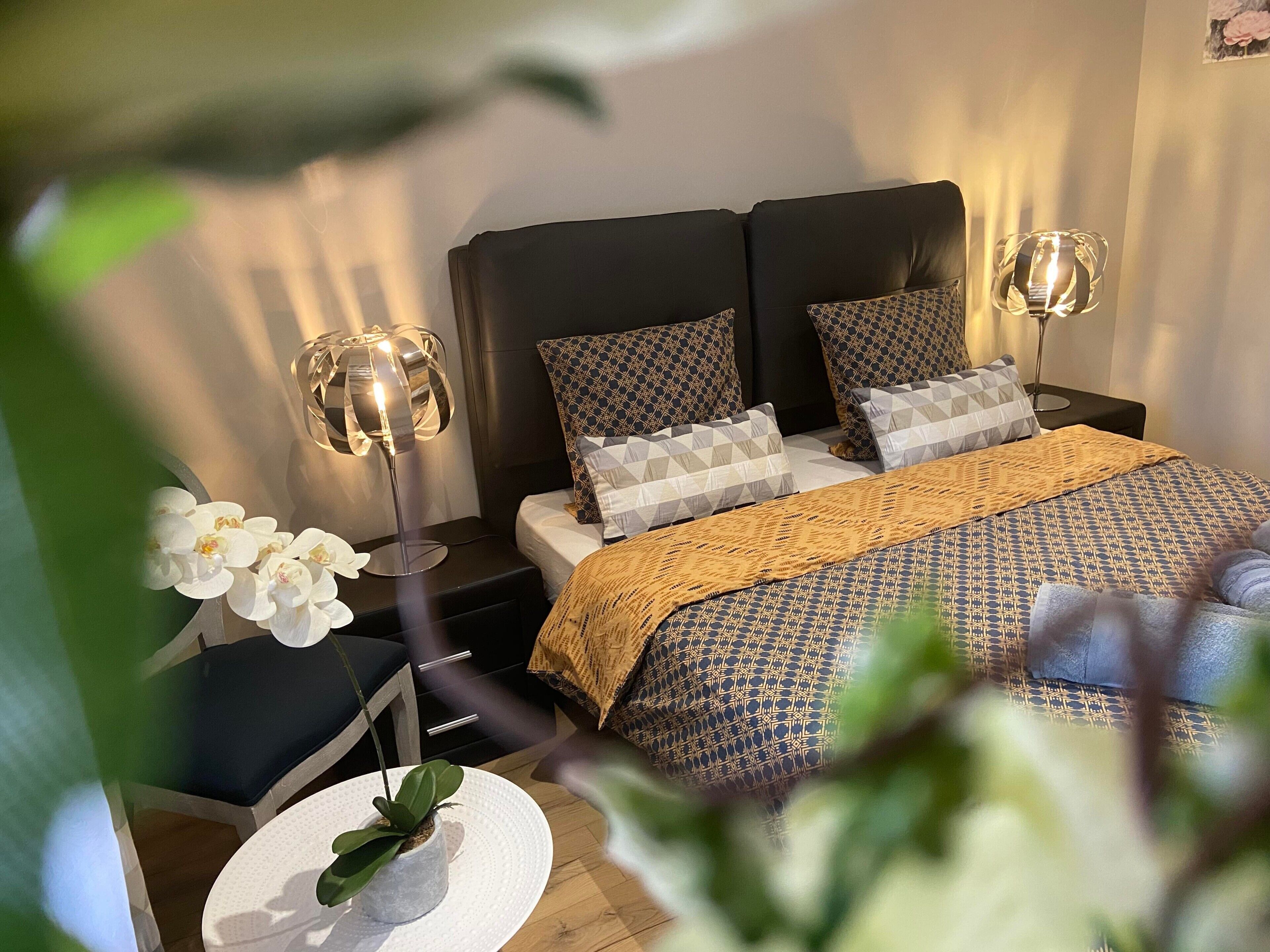 Een slaapkamer met een bed, twee nachtkastjes, een lamp, een potplant en een kunstwerk aan de muur.