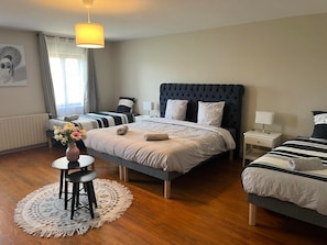 Ein Hotelzimmer mit einem großen Bett, einem kleineren Bett, einem Nachttisch mit Lampe, einem runden Tisch mit Blumenvase und einer Pendelleuchte.