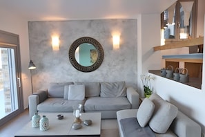 A modern living room with a grey sofa, a coffee table with decorative items, a round mirror, and a wooden shelf with storage baskets. - Villa 'Lina Syros' with Sea View, Wi-Fi and Air Conditioning (Syros)