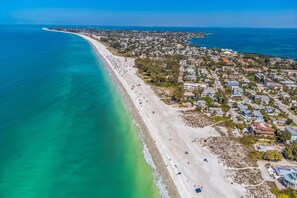 Vista aérea de uma vila costeira com uma extensa praia de areia, águas turquesas cristalinas e edifícios residenciais.