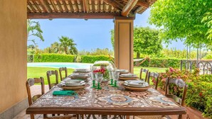 An outdoor dining area with a patterned tablecloth, wicker chairs, and a view of a swimming pool and greenery.