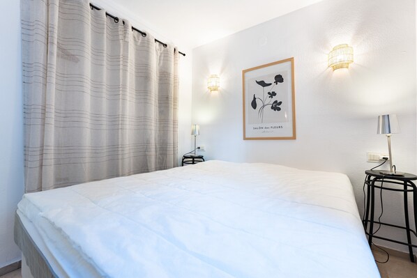 A bedroom with a bed, curtains, a wall-mounted light, a framed picture, and a small table with a lamp.