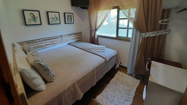 A bedroom with a bed, a window with curtains, a mounted TV, and framed pictures on the wall. - Pousada Sítio Bicho do Mato  (Tijucas)
