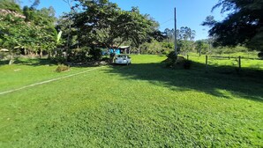 A grassy area with a white car parked near a tree, surrounded by greenery and a clear blue sky. - Pousada Sítio Bicho do Mato  (Tijucas)