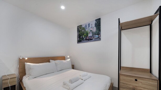 A hotel room with a wooden bed, white bedding, a bedside lamp, a small wooden nightstand, a wall-mounted mirror, and a cityscape picture. - Cosy Studio (Redfern)