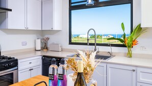 A modern kitchen with white cabinets, a stove, and a dishwasher. There is a window with a view of the sea and a plant on the counter. - Luxury Cliffside Oceanview Villa with Private Pool and Rooftop. Accommodates 7. (Isabela)