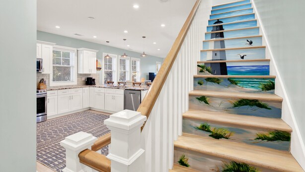 Une cuisine moderne avec des meubles blancs, un îlot central et un escalier orné d’une fresque représentant un paysage marin.