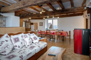 Un salon chaleureux avec un plafond en bois, un canapé agrémenté de coussins à motifs, une table à manger avec des chaises, et un réfrigérateur rouge et noir.