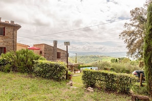 Un bâtiment en pierre avec un toit rouge, un jardin bordé de haies et une vue sur des collines lointaines.