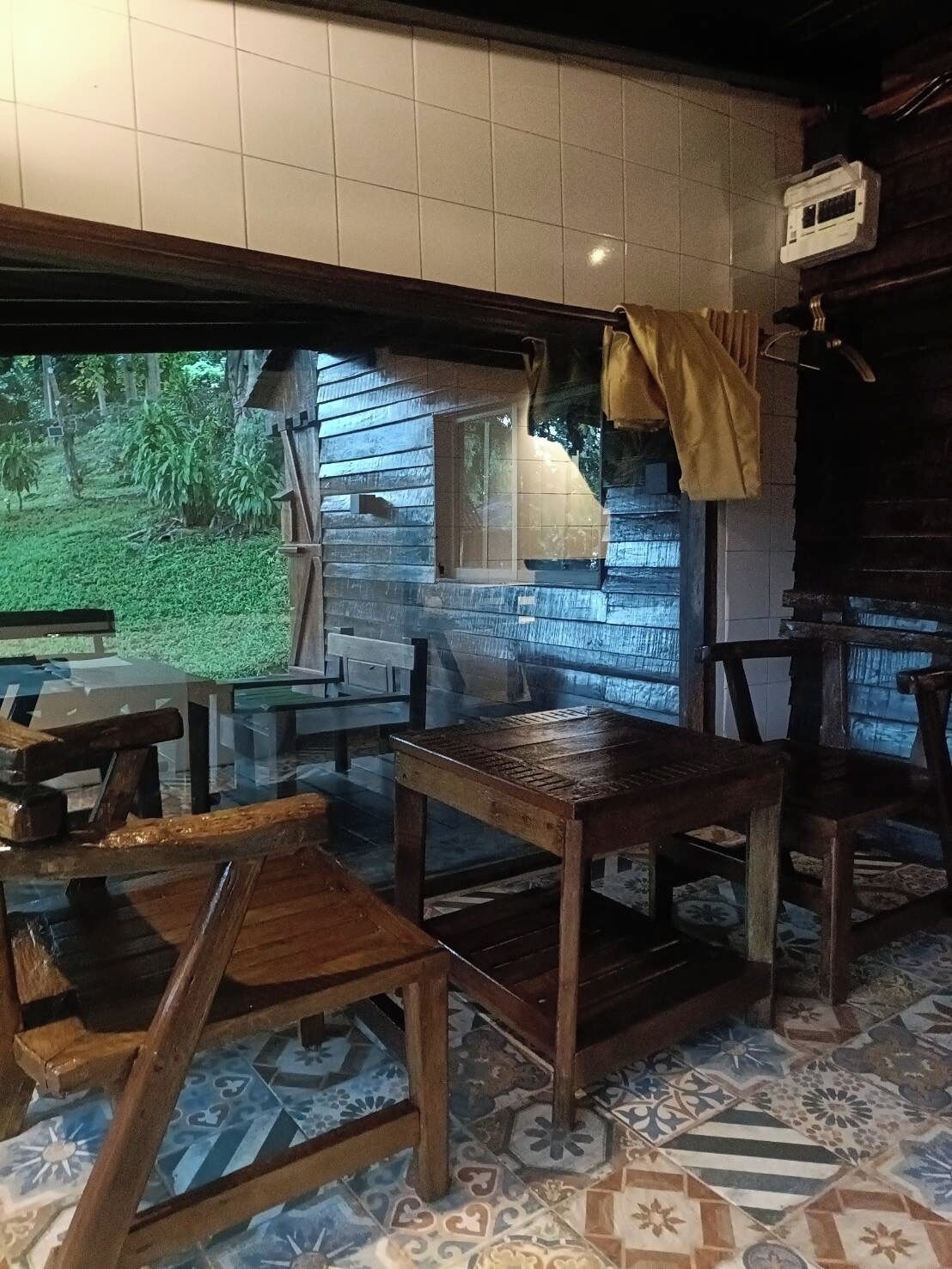 A rustic wooden dining area with a glass table and chairs, a tiled floor with a geometric pattern, and a view of greenery outside.