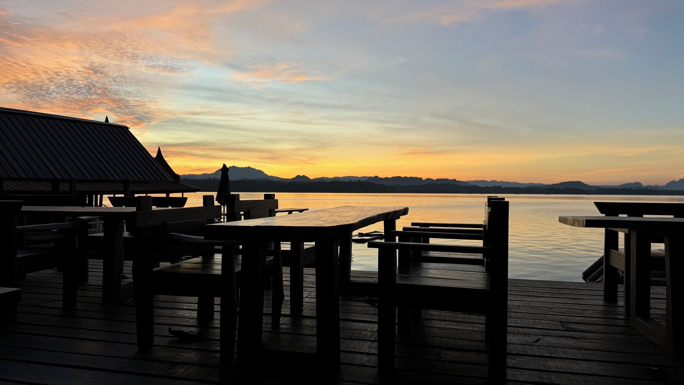 Eine Holzterrasse mit Tischen und Stühlen, die auf ein Gewässer im Sonnenuntergang blickt.