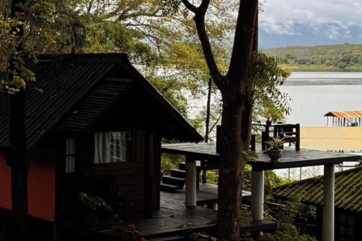 A cabin with a wooden deck overlooking a lake, surrounded by trees.