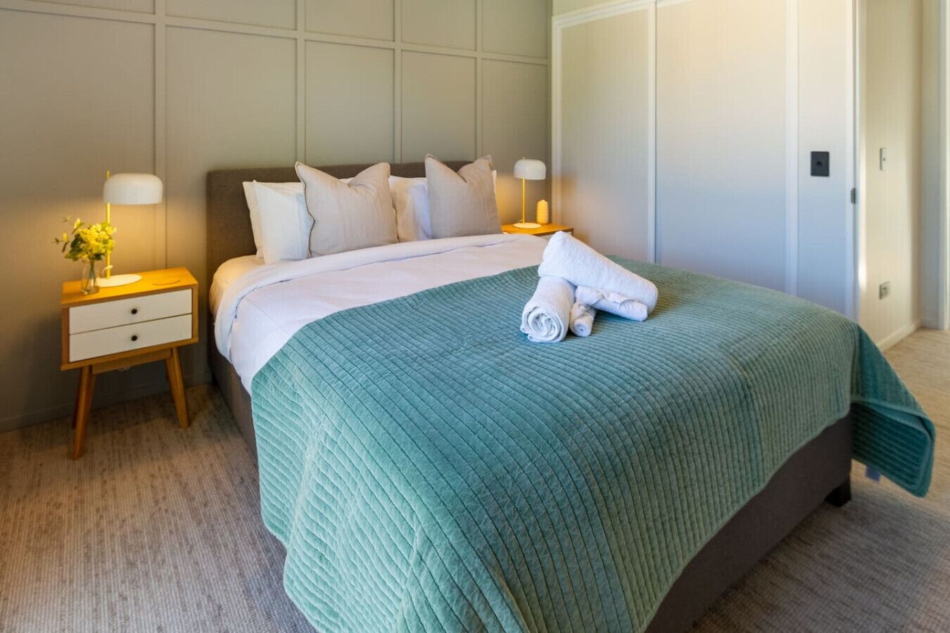 A neatly made bed with a teal bedspread, two bedside tables with lamps, and a door to the right.