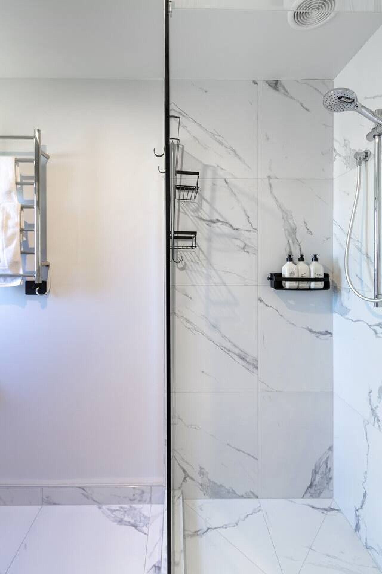 A modern bathroom with a glass shower enclosure, marble tiles, a towel rack, and a soap dispenser shelf.