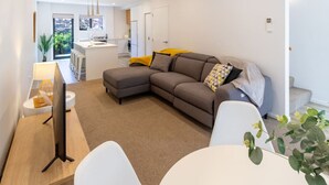 A modern living room with a grey sectional sofa, a white coffee table, and a TV on a wooden stand. There is a kitchen in the background with white cabinets and a yellow towel on the counter.