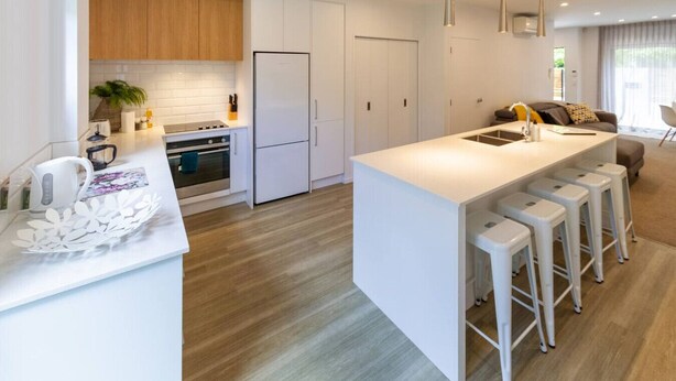 A modern kitchen with white cabinetry, a central island with bar stools, and a dining area with a sofa and table.