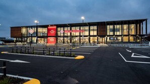 A modern building with a large glass facade, a prominent red banner, and a parking lot with directional arrows.