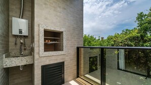 A modern bathroom with a built-in fireplace, a water heater, and a glass-enclosed balcony with a view of greenery. - Apartamento com 02 Dormitórios em Canto Grande (Bombinhas)