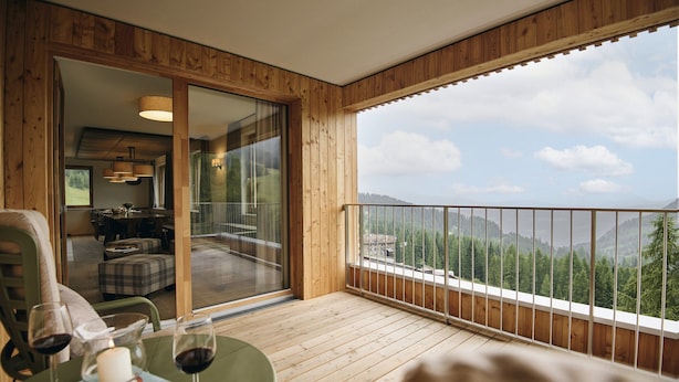 Un balcon en bois offrant une vue sur les montagnes et les arbres, une porte vitrée donnant sur un coin repas, et une table avec des verres à vin et un livre.