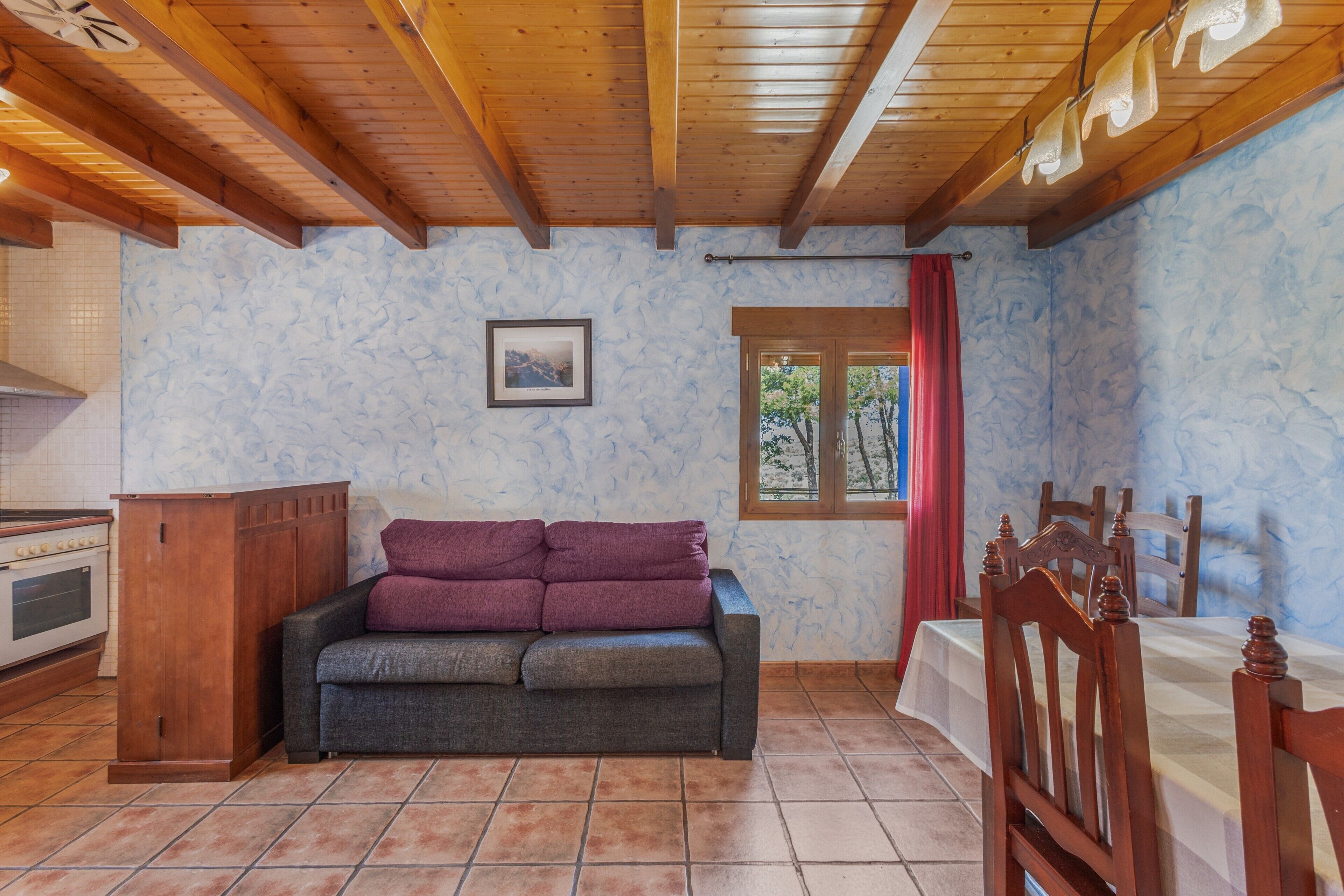Une pièce avec un plafond en bois, un canapé, une table à manger avec des chaises, une fenêtre donnant sur des arbres et un coin cuisine avec une cuisinière.
