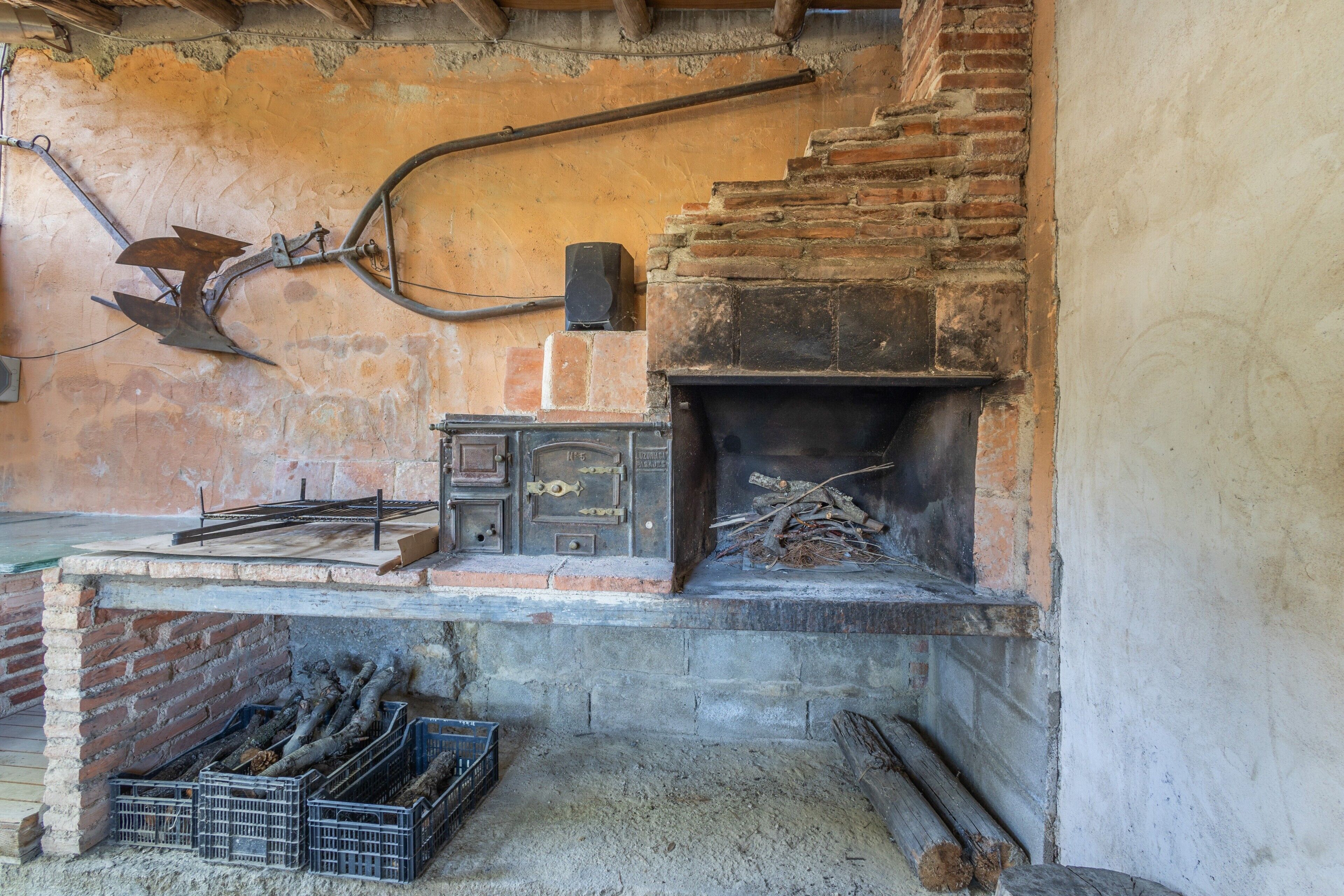 Une cheminée en briques au style rustique, avec des bûches, un poêle en métal et une étagère en bois où sont rangés des outils.