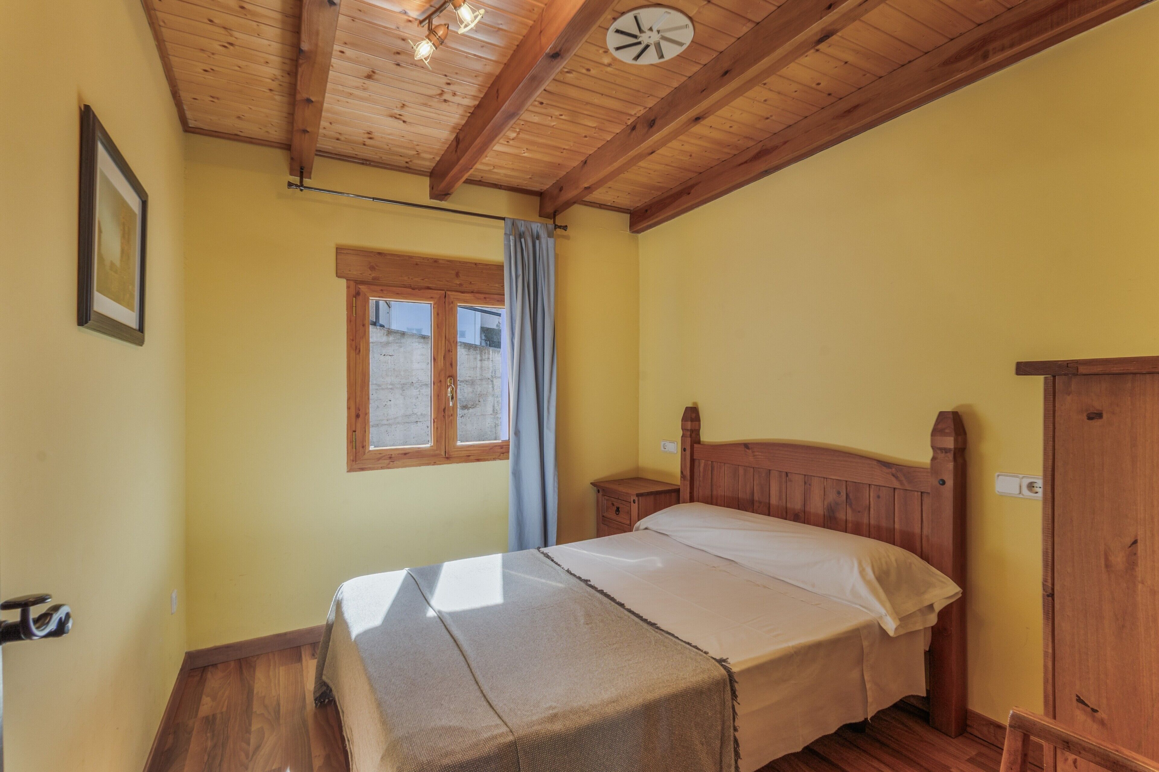 A bedroom with a wooden ceiling, a bed with a wooden headboard, a nightstand, a window with a curtain, and a wall clock.