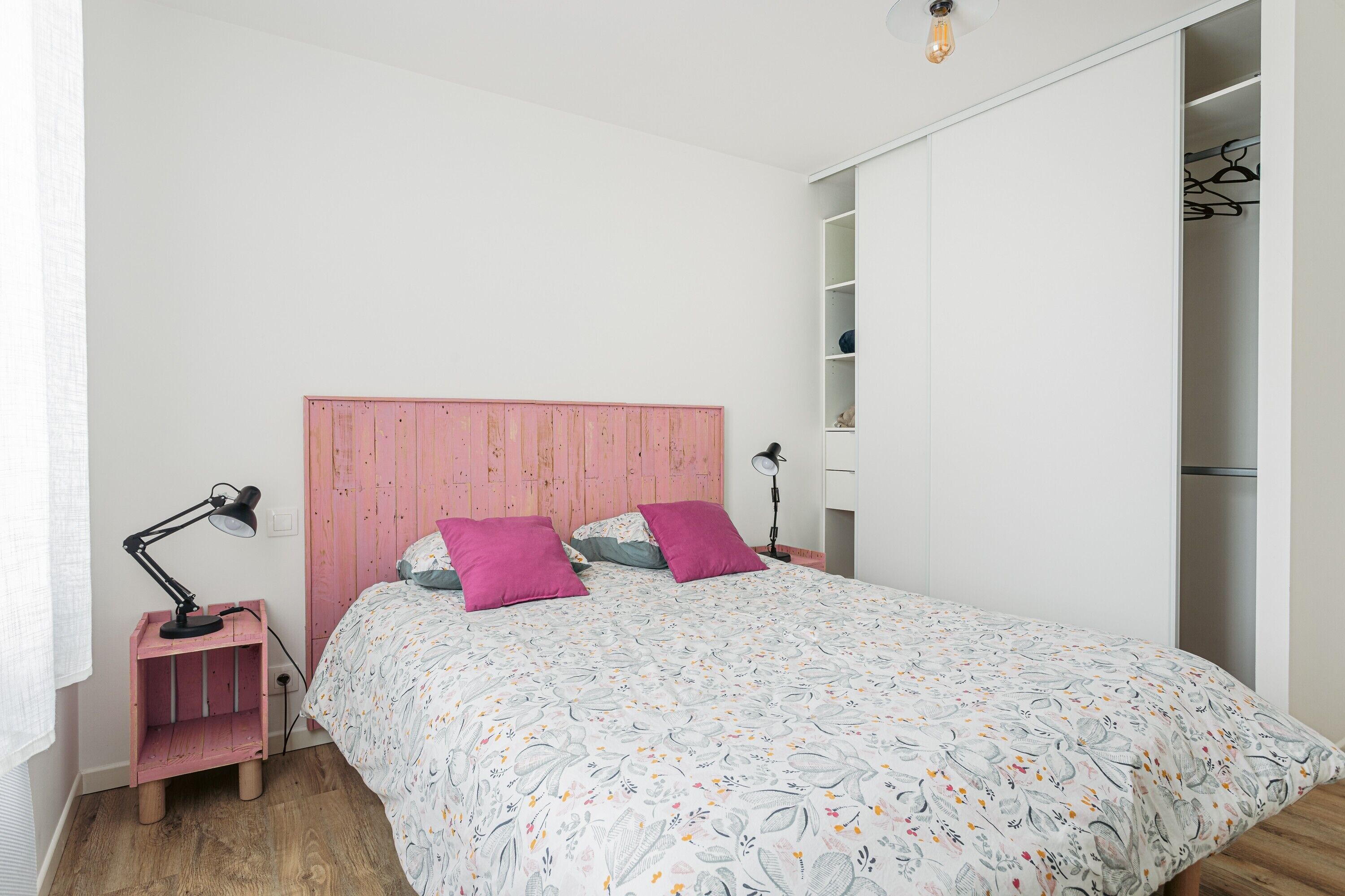 Een slaapkamer met een bed, een roze hoofdbord, twee roze kussens, een bedlampje en een garderobe.