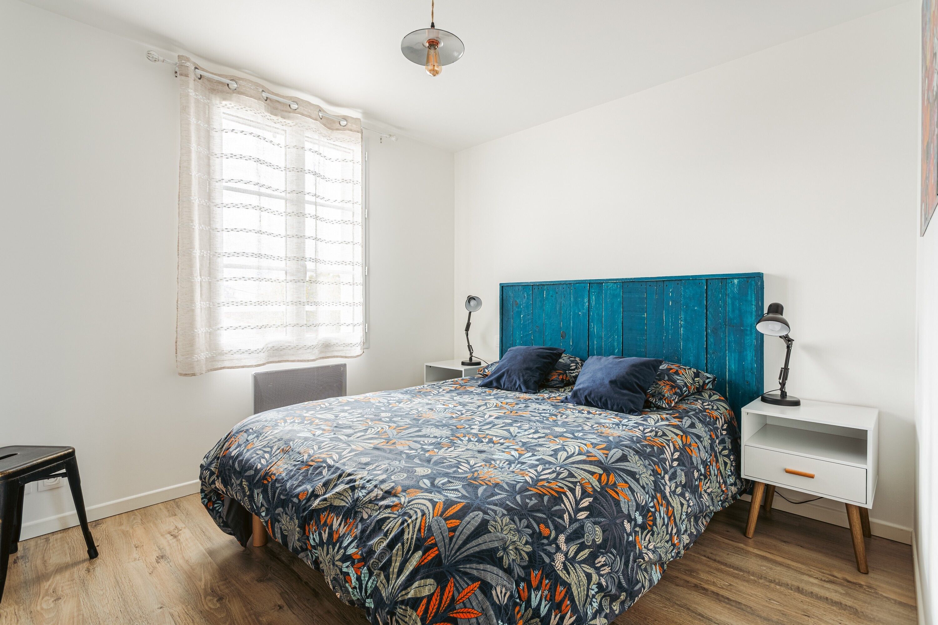 Un dormitorio con cama, mesita de noche, lámpara, silla y ventana con cortinas.