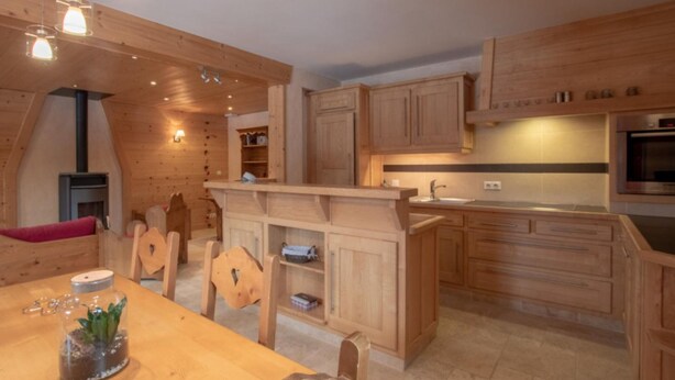 A wooden interior with a dining table, chairs, a kitchen area with wooden cabinets, a built-in oven, and pendant lighting. - Apartment 'La Grange - 4 Pers' with Mountain View, Private Garden and Wi-Fi (Taninges)