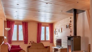 A cozy room with a wooden ceiling, a sofa, a chair, a small table, a fireplace, and a window with curtains. - Apartment 'La Grange - 4 Pers' with Mountain View, Private Garden and Wi-Fi (Taninges)