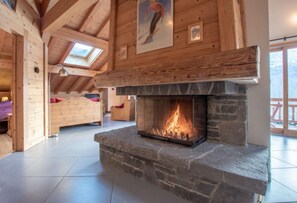 Interior de una cabaña de troncos con chimenea de piedra, vigas de madera, una cama y un tragaluz.