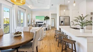 Cozinha moderna com ilha central, sala de jantar com mesa de madeira e sala de estar com sofá e televisão.